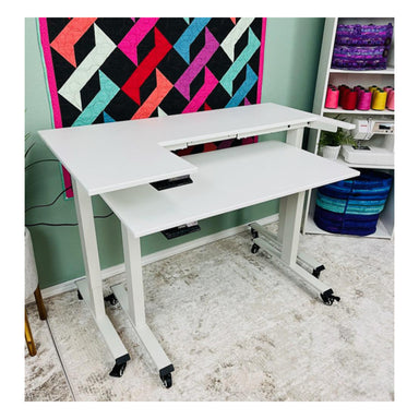 A white Classi Creations Dual Tier Adjustable Quilting and Sewing Table on wheels is pictured in a colorful room. Behind the desk, there is a quilt with a geometric pattern of black, pink, green, and red. Shelves to the right hold rainbow-colored thread and folded fabric. The floor is carpeted.