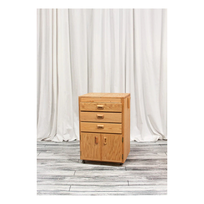 A compact, four-drawer BEST Sitha Taboret stands on a light-toned wooden floor against a background of white, floor-length curtains. Perfect for art supplies storage, the taboret has three small upper drawers and one larger drawer at the bottom, all with simple wooden handles.