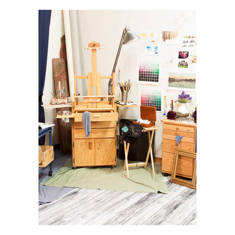 An artist's studio with a wooden easel, paintbrushes, a palette, and art supplies on a BEST Sitha Taboret from BEST. Numerous artworks, photos, and color swatches hang on the wall. The room has a cozy, creative atmosphere with a lamp illuminating the workspace.