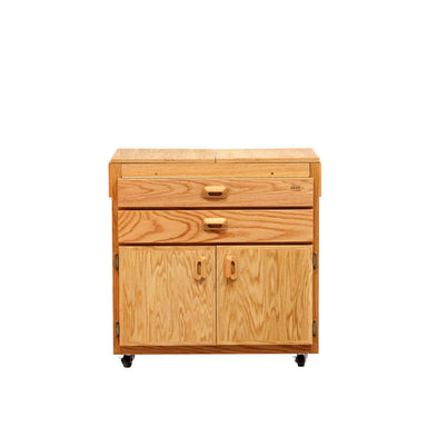 The BEST Caitlin Taboret by BEST is a wooden kitchen cart with a natural finish, featuring a top drawer, a middle compartment with a smaller drawer, and a double-door cabinet at the bottom. The cart is on wheels for mobility and has simple, rectangular handles. Inspired by an oak taboret design, it brings both elegance and functionality to your kitchen.