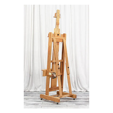 A tall, sturdy BEST Abiquiu Easel w/ Optional Tray designed for holding canvases, standing on a wooden floor with a white curtain backdrop. The easel features adjustable height and support mechanisms and is equipped with wheels for mobility.