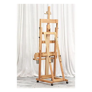 A single-masted easel stands against a backdrop of white, draped fabric. The BEST Classic Santa Fe I Easel by BEST is adjustable, featuring various knobs and mechanisms to hold and secure a canvas. It includes a painting tray for convenience and has a sturdy base with wheels, making it easy to move around.