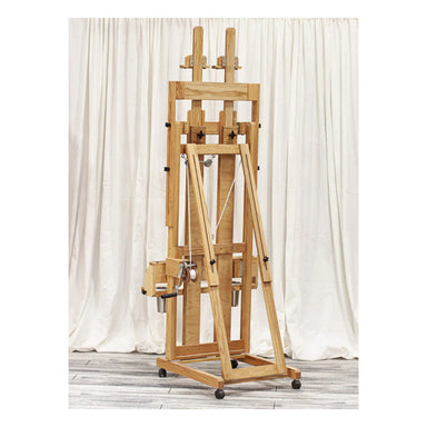 The BEST Classic Santa Fe II Easel, reminiscent of a double-masted H-frame, stands against a plain white curtain backdrop. The easel boasts multiple adjustable components, including a shelf and clamps to hold canvases, and is equipped with wheels for easy mobility. The floor beneath is light-colored wood.