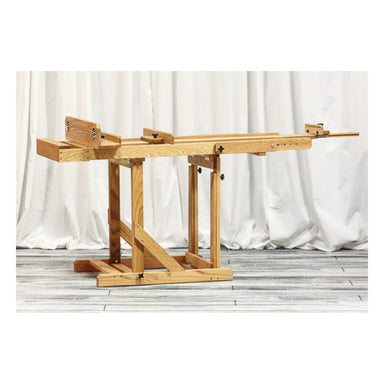 A BEST European Easel by BEST is placed on a wooden stand with a white curtain background and a wooden floor. The easel features a detailed construction with angled supports and various mechanisms for adjusting, reminiscent of the craftsmanship seen in an adjustable crossbow.
