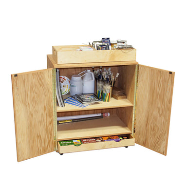An open oak art cart with two doors reveals shelves stocked with various art materials including paintbrushes, books, sketchpads, paints, and other supplies. A small wooden box on top holds additional tools and paint containers, making it the BEST Art Cart by BEST for any artist.